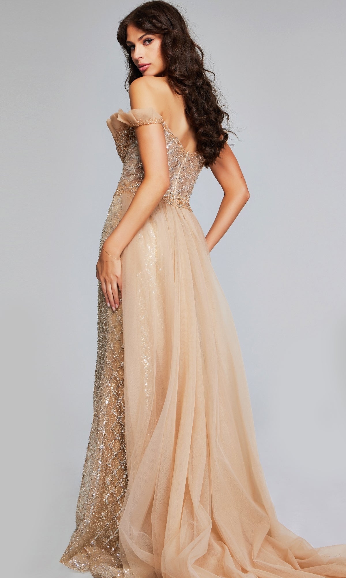 A woman poses with her back slightly turned, wearing the Jovani 39389โa long formal off-the-shoulder mermaid dress featuring floor-length sequins and a sheer overlay.